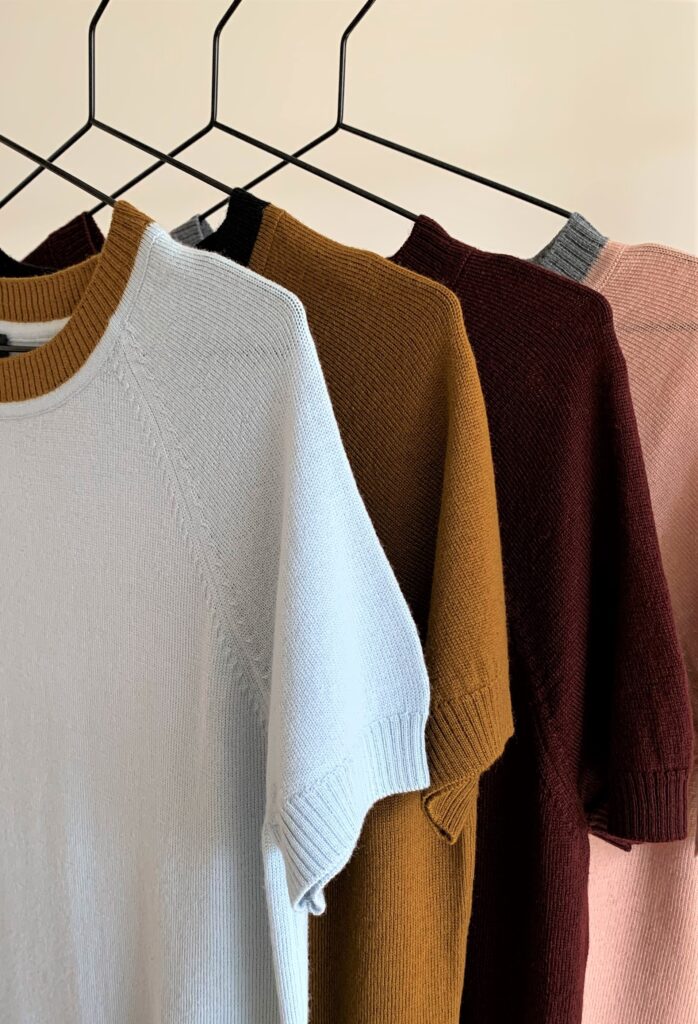 Colorful knit sweaters hang on a hanger. About us, Hanne Linding.